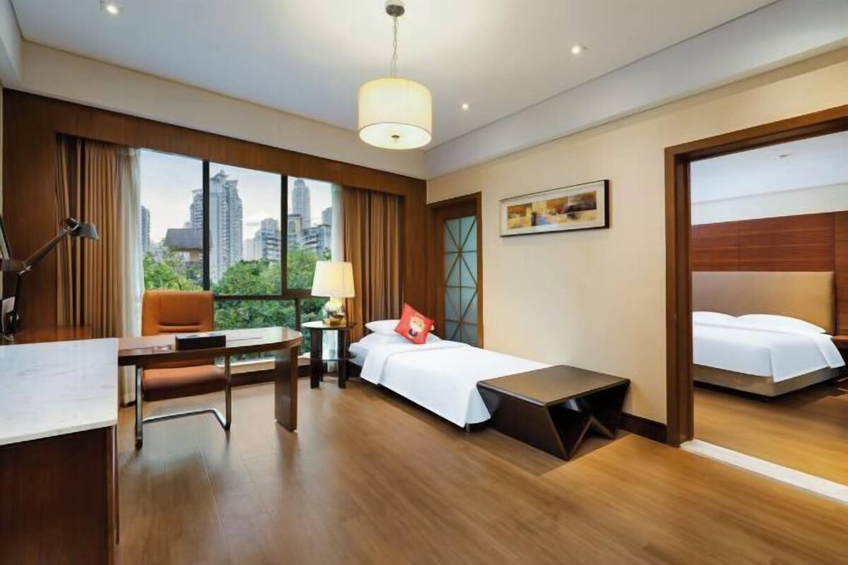A hotel room with a desk, a bed, a chair, a lamp, and a view of the city.