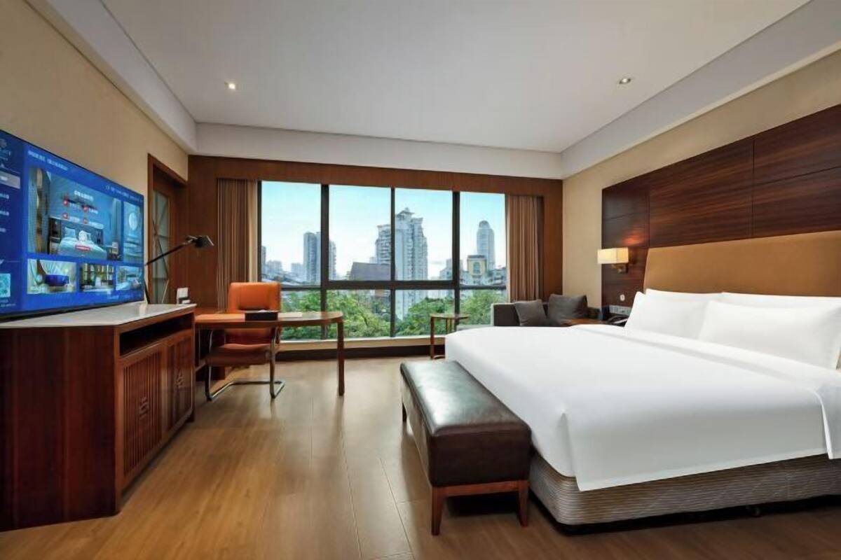 A hotel room with a large bed, a desk with a computer, a chair, and a view of the city skyline through large windows.