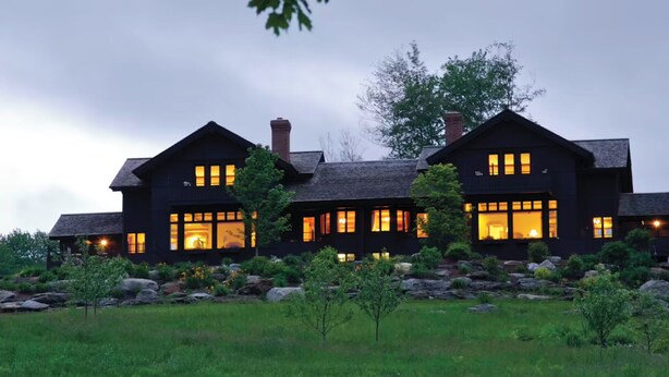 A large, dark-colored house with multiple windows illuminated from the inside, surrounded by a well-maintained lawn and rocks.