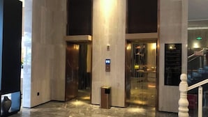 En moderne lobby med marmorgulv, en central elevator og en trappe med hvidt gelænder.