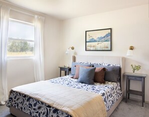Une chambre à coucher avec un lit, une fenêtre avec des rideaux, une table de chevet avec une lampe et un tableau accroché au mur.