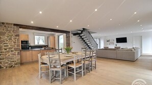 A modern kitchen with a stone accent wall, a dining area with a wooden table and chairs, a staircase, and a living area with a sofa and a TV.
