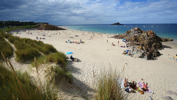 A beach with people sunbathing and swimming, a rocky outcrop, and boats in the water.