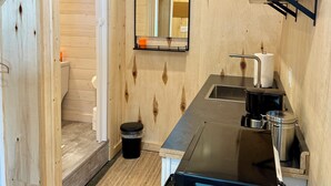 Un baño moderno y compacto con paredes de madera y ducha integrada. Incluye un lavamanos, un espejo y una estantería con un cuenco.