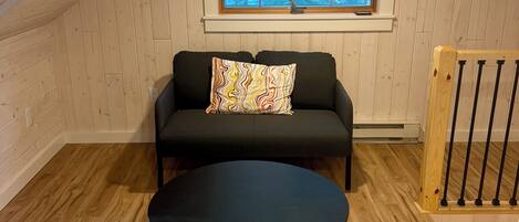 Una habitación acogedora en el ático con suelo de madera, un sofá negro con un cojín de colores, una mesa redonda negra y una ventana con vistas a árboles.