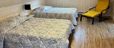 Une chambre mansardée chaleureuse avec un plancher en bois, deux lits avec une literie à motifs, une chaise jaune, une petite fenêtre et une lampe de chevet.