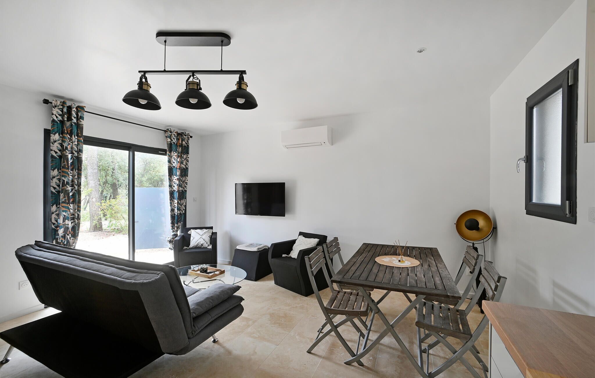 A modern living room with a sofa, a dining table with chairs, a flat-screen TV, and a ceiling light fixture.