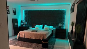 A modern bedroom with a large bed, bedside tables, a nightstand, and a wall-mounted shelf. - Sejour's Penthouse (Santo Domingo Este)