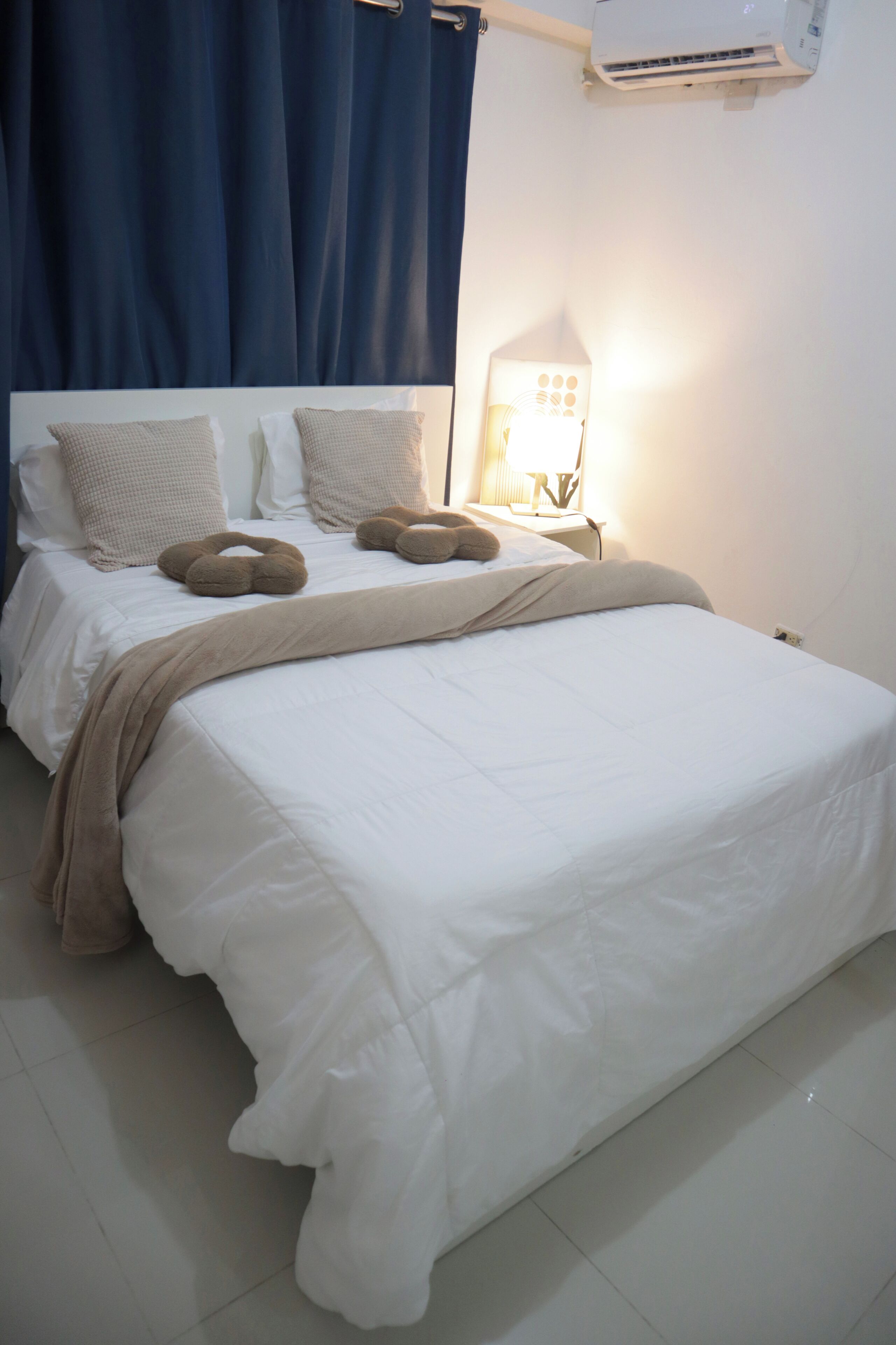 A neatly made bed with a white comforter and two brown pillows, a bedside lamp, and a wall-mounted air conditioner.