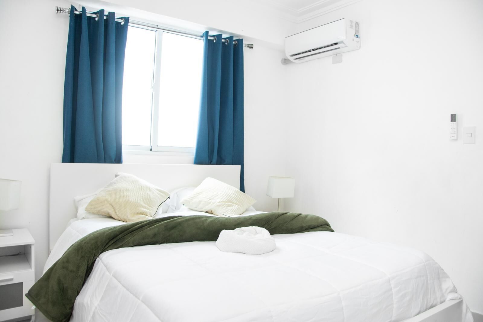 Ein ordentlich bezogenes Bett mit weißer Bettwäsche und einer grünen Decke, ein Nachttisch mit Lampe und ein Fenster mit blauen Vorhängen.
