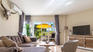 Un salon moderne avec un canapé, un pouf, une télévision et un coin repas agrémenté d’un parasol jaune.