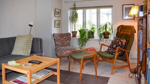Ein gemütliches Wohnzimmer mit grauem Sofa, einem gemusterten Sessel und einem Couchtisch mit Büchern und einer Fernbedienung.
