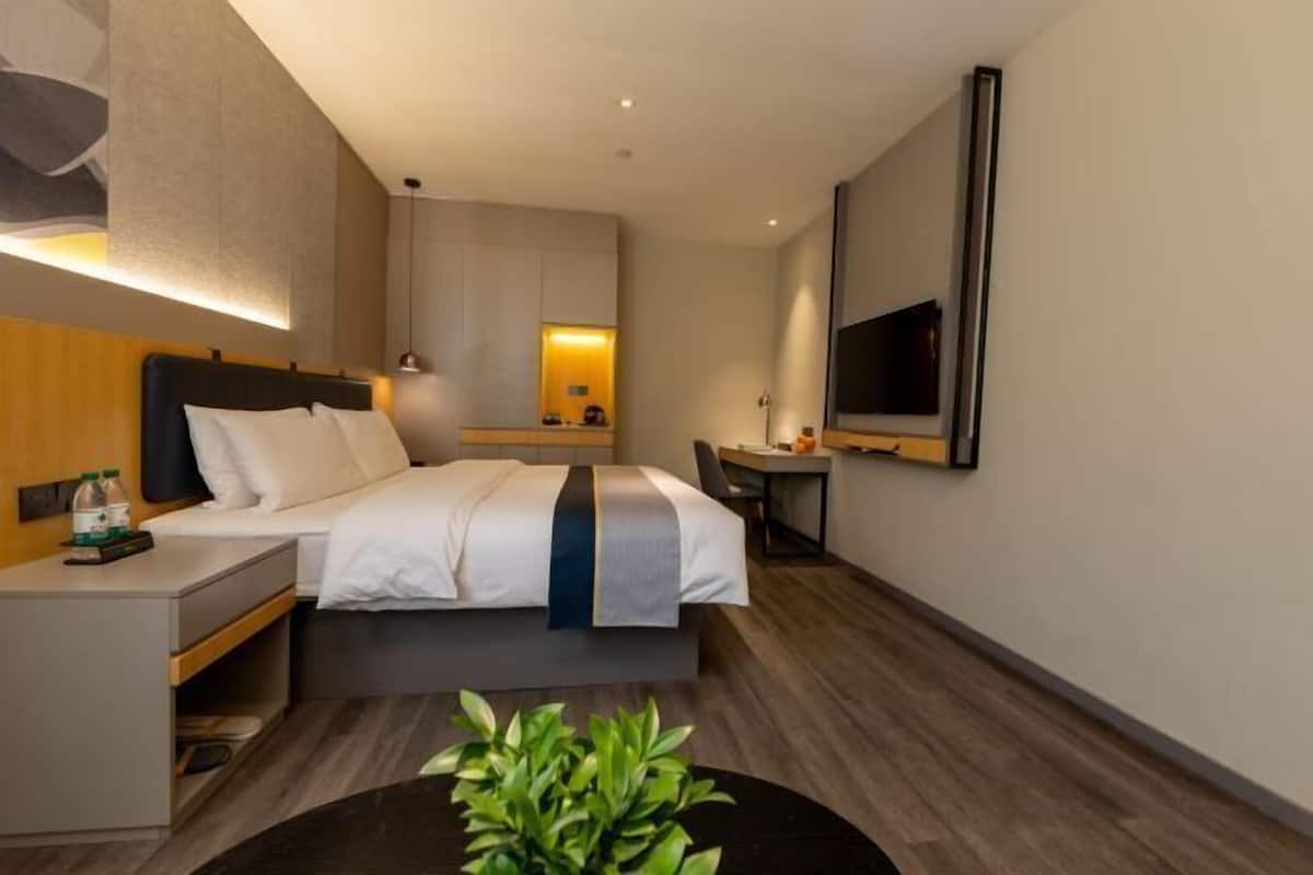 Moderni hotellihuone, jossa on sänky, yöpöytä, televisio ja pieni työpöytä tuolilla.