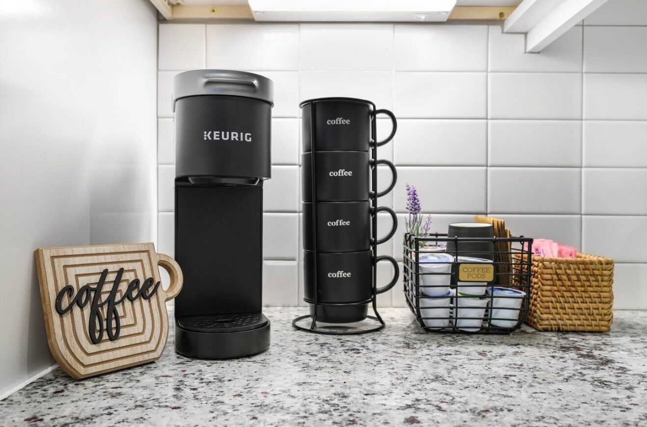 A kitchen with a coffee maker, a stack of coffee mugs, a wooden coffee sign, and a basket of coffee pods.