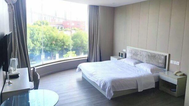 A hotel room with a bed, bedside tables, a desk with a chair, a TV, and a large window offering a view of trees and buildings. - Changwei Hotel (Shanghai)