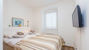 Une chambre à coucher avec un lit, un téléviseur fixé au mur, une fenêtre avec des stores et un tableau encadré accroché au mur.