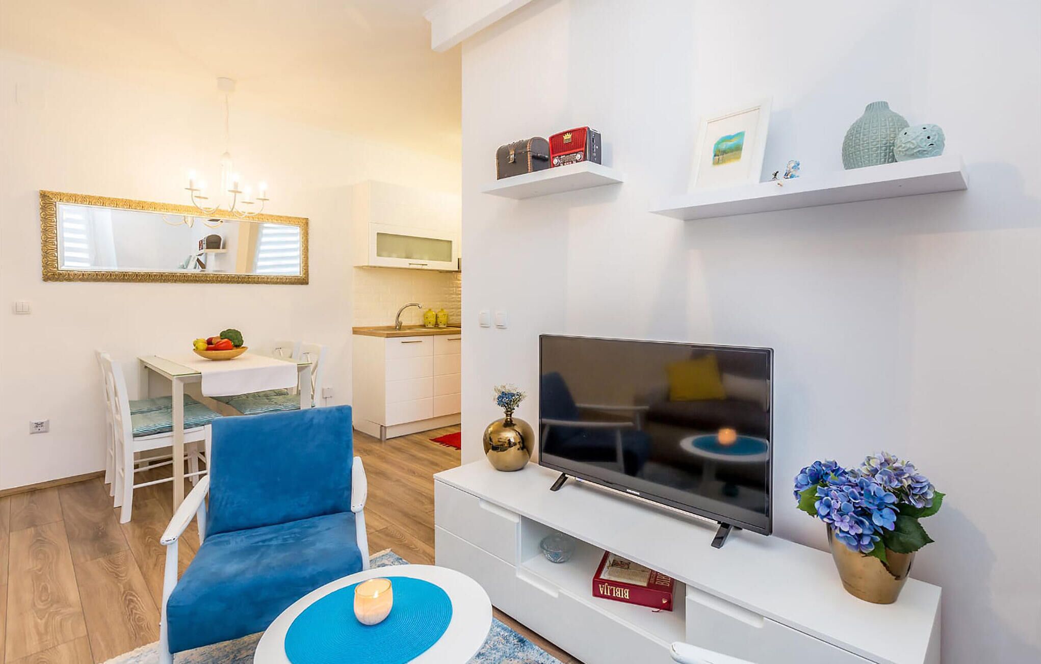 Un salon moderne avec un téléviseur, un canapé bleu, un coin repas avec une table et des chaises, et une cuisine en arrière-plan.