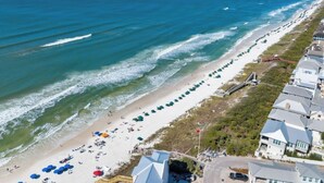 Ein Küstenstrand mit einer langen Reihe von Strandsonnenschirmen und Liegestühlen, einer Holzpromenade und einer Reihe Häuser mit geneigten Dächern.