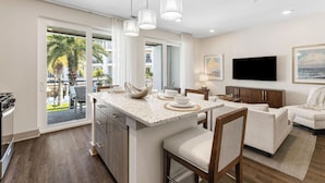 Eine moderne Küche mit Kochinsel, ein Essbereich mit weißem Tisch und Stühlen, ein Wohnzimmer mit Sofa und Flachbildfernseher sowie ein Balkon mit Blick auf Palmen.