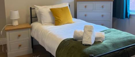A neatly made bed with a green blanket and two towels, a white bedside table with a lamp, a yellow pillow, and a white dresser with drawers.