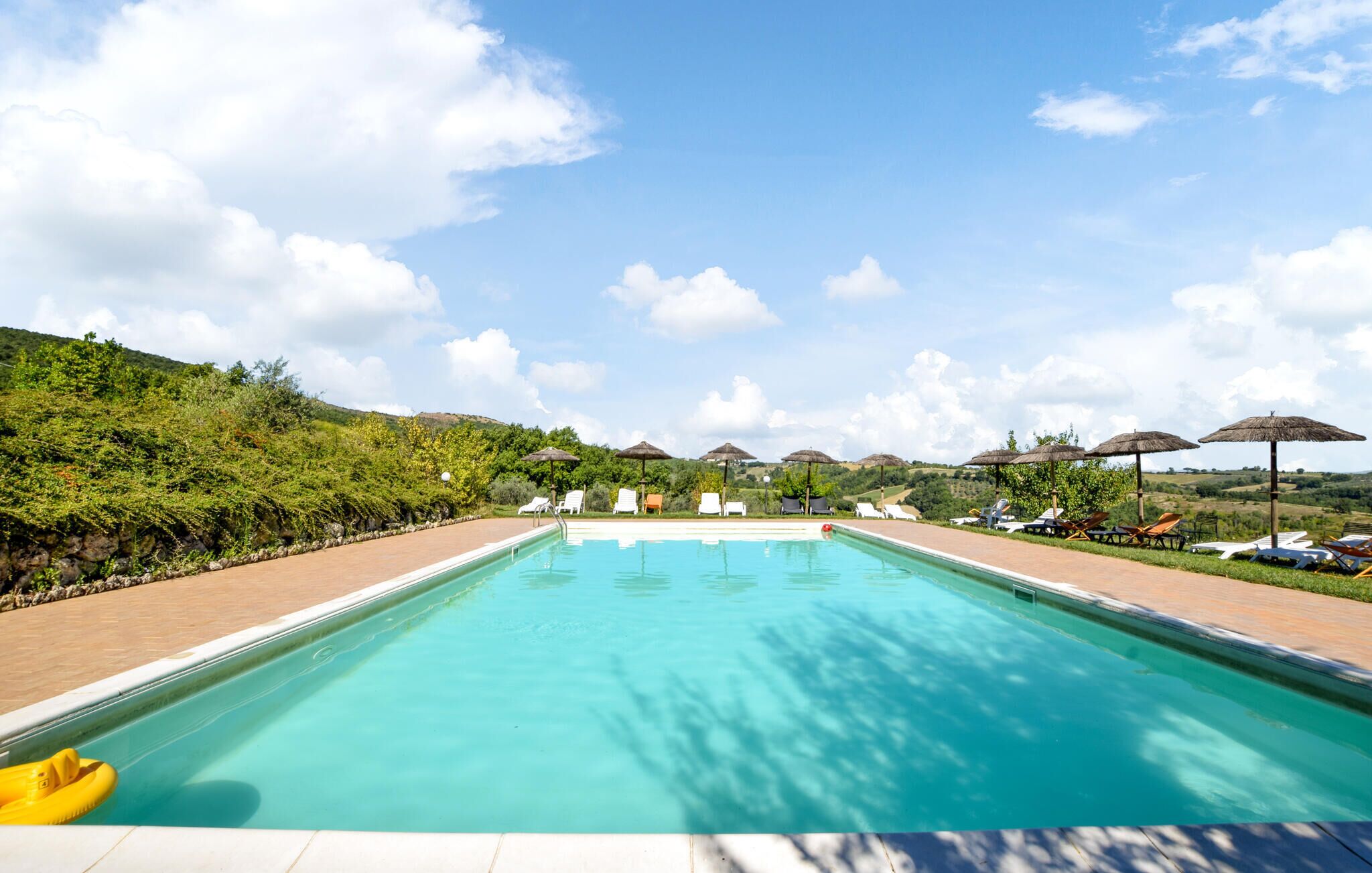 A swimming pool with lounge chairs and umbrellas, surrounded by greenery and hills.