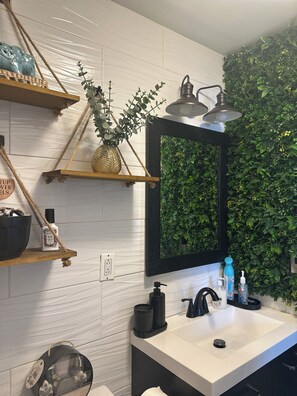 Une salle de bain moderne avec un mur recouvert de carreaux blancs, un miroir à cadre noir, un mur végétal, et un lavabo blanc agrémenté de divers produits de toilette.