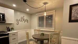 Un comedor con una mesa redonda, cuatro sillas, una lámpara de pared y un ventanal con persianas.