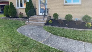 Una casa de dos pisos con porche delantero, fachada de ladrillo y un pequeño jardín con diversas plantas y flores.