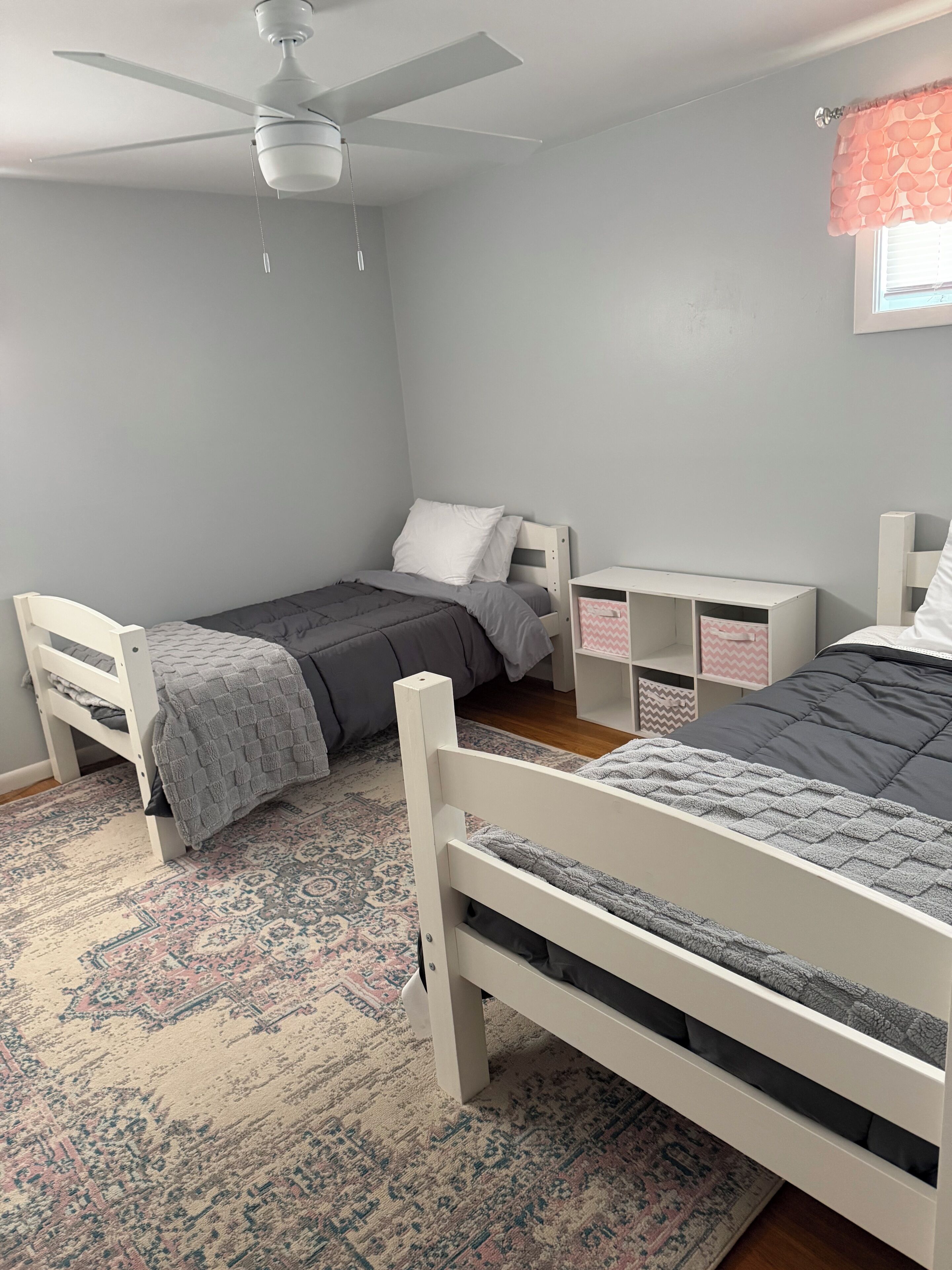 Un dormitorio con dos camas individuales, ventilador de techo, ventana con cortina rosa y alfombra estampada.
