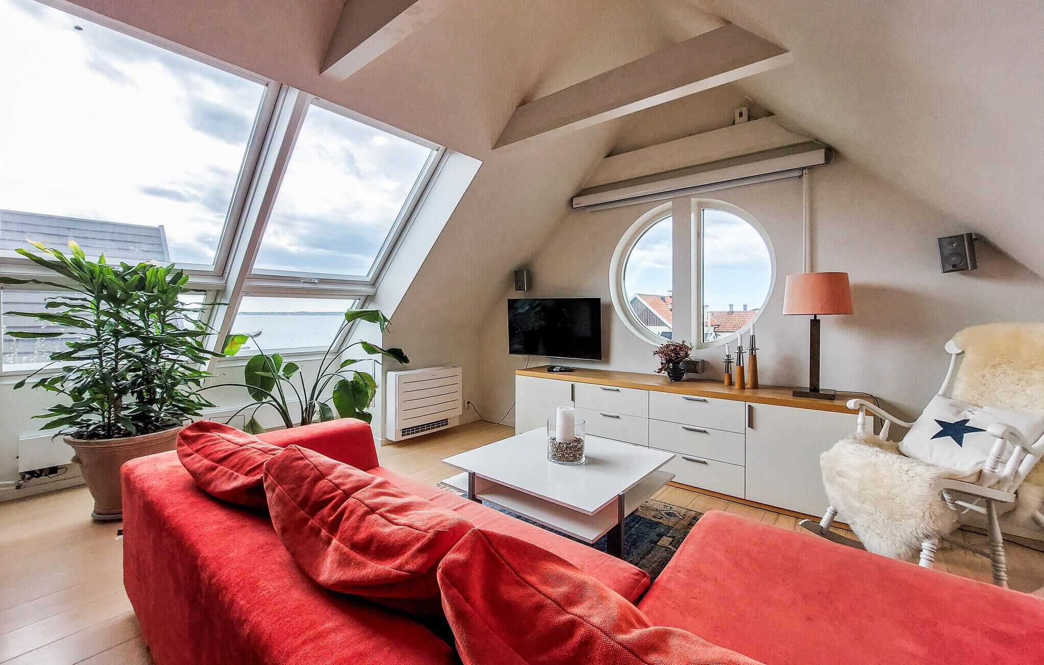Ett modernt sovrum i loftstil med ett stort takfönster, en tv, en rund spegel och en mysig röd soffa.