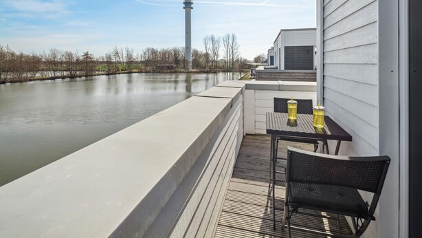 Un balcon avec vue sur une rivière, un phare visible au loin et une table avec des chaises.