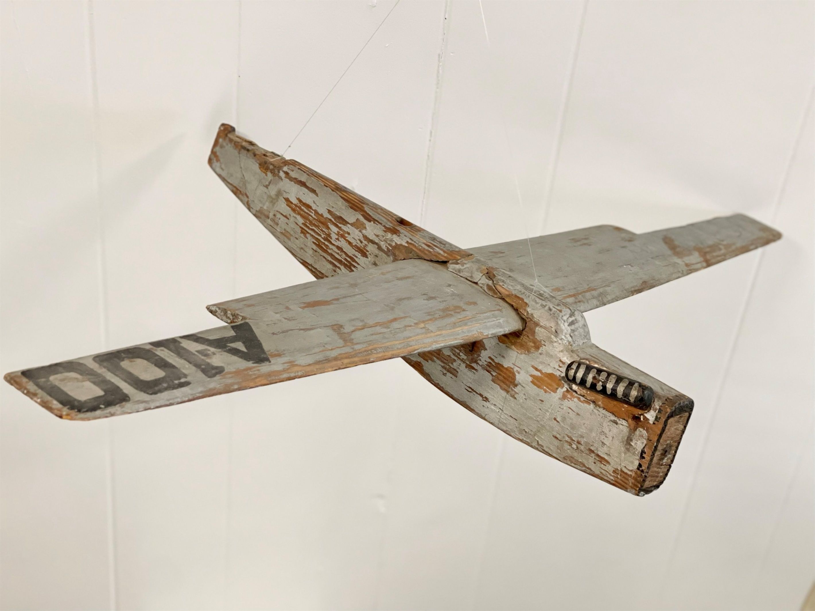 Un modèle d’avion en bois, d’aspect rustique et patiné par les intempéries, avec une hélice bien visible, fixé à un mur blanc.