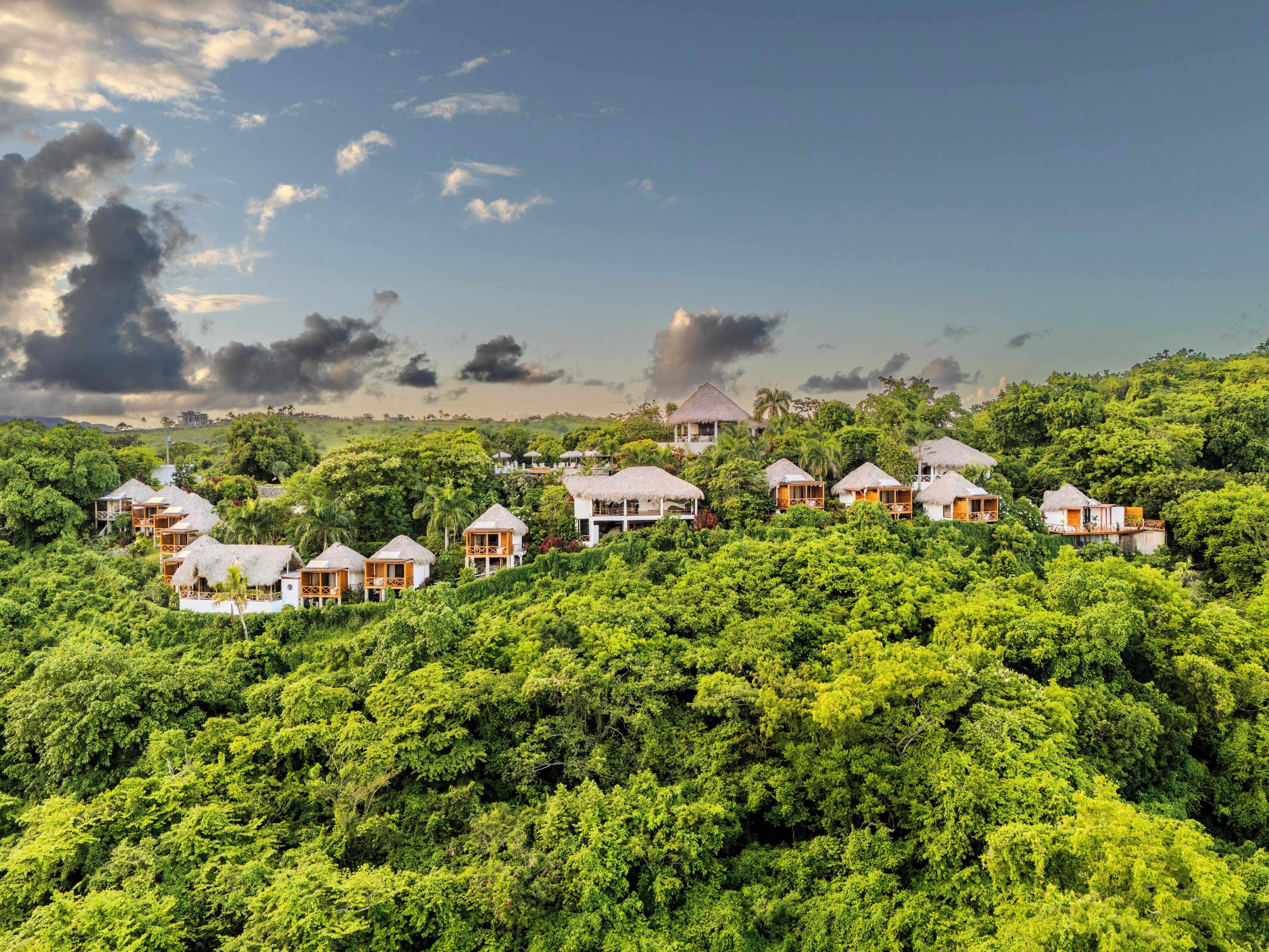 A resort with thatched-roof bungalows nestled among lush greenery, overlooking a hilly landscape.