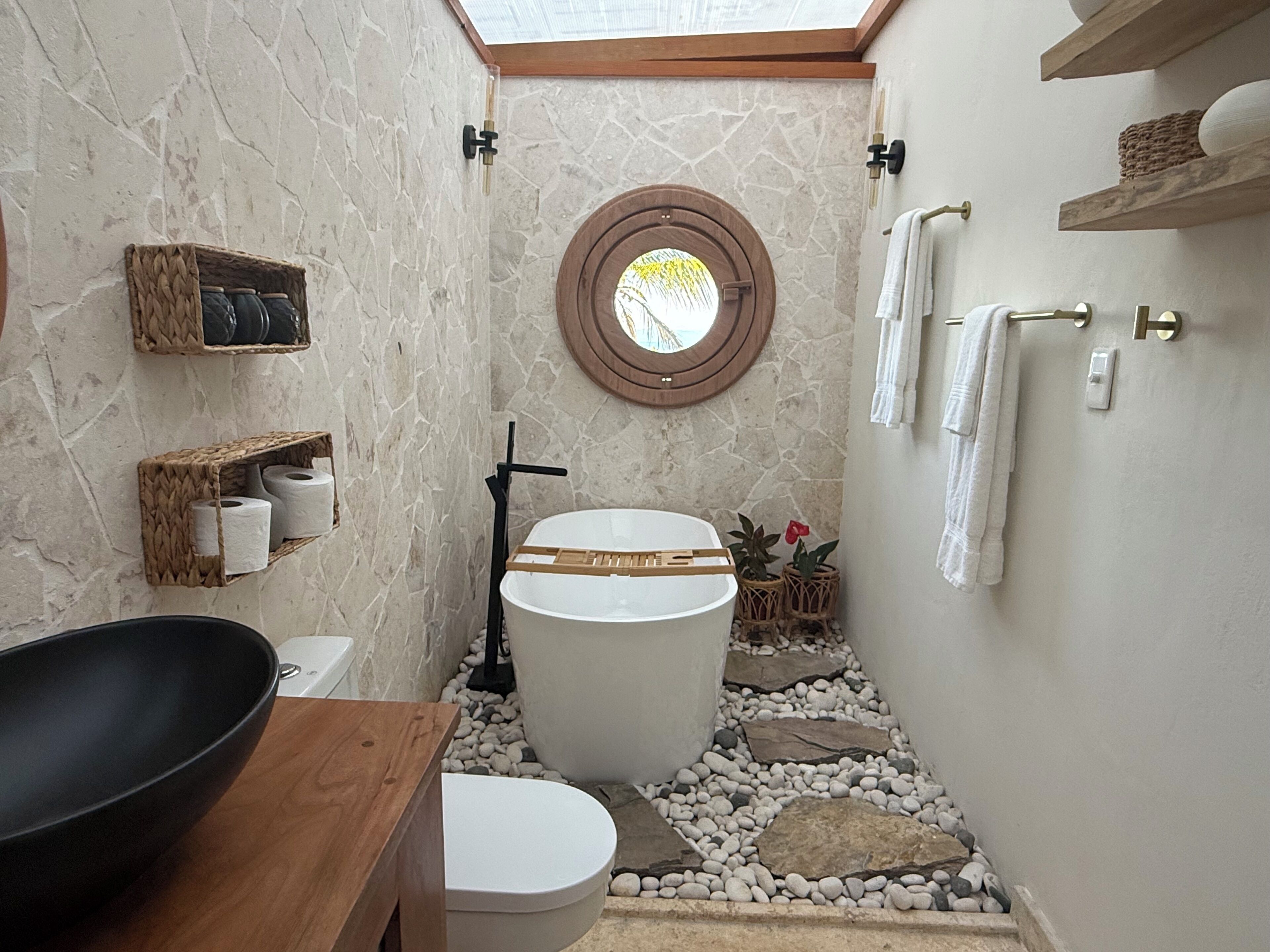 Un baño con pared de piedra, tocador de madera, un lavabo negro grande, inodoro y ducha con puerta de madera, además de un tragaluz.