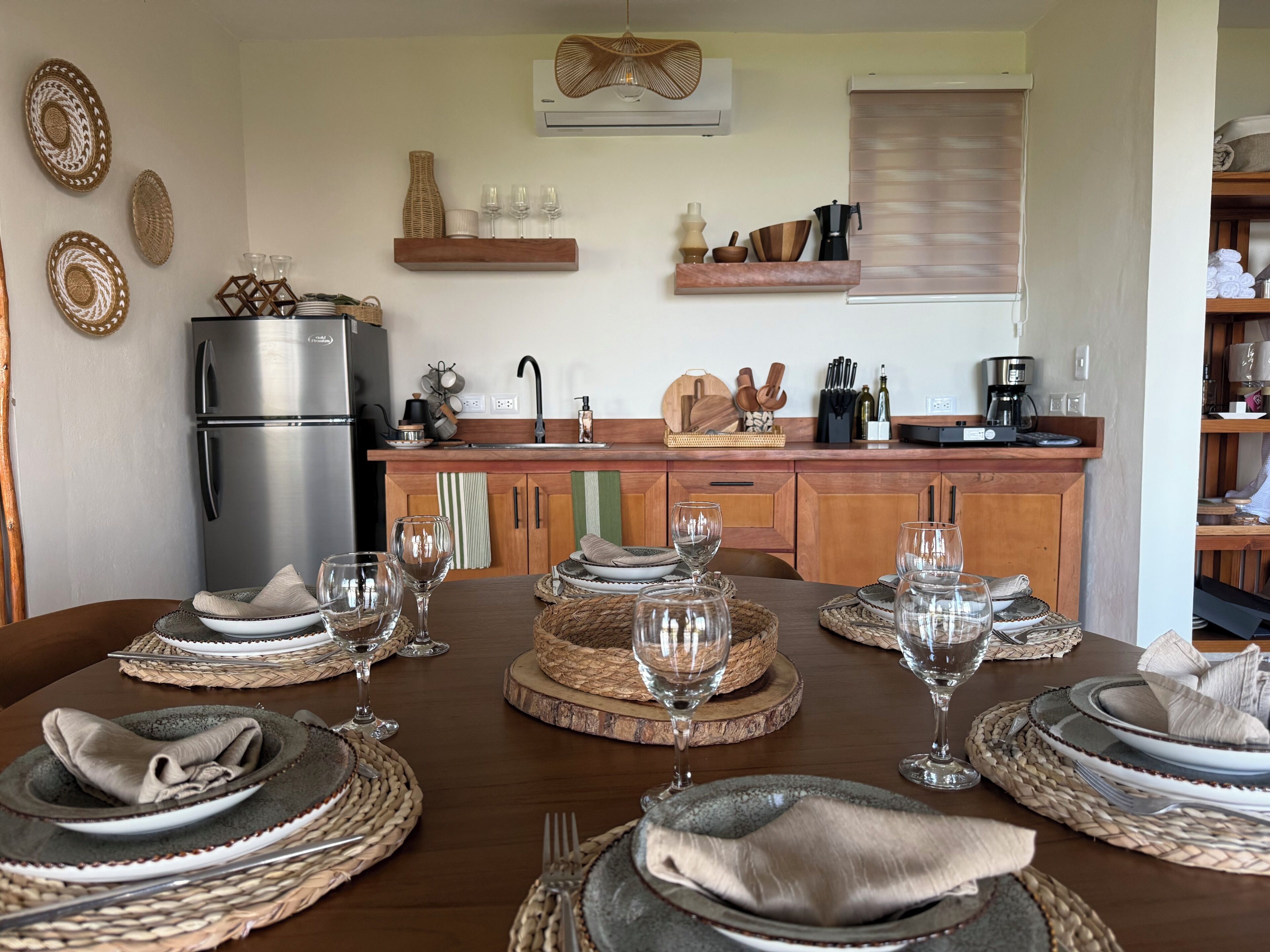 A dining area with a wooden table set for four, a stainless steel refrigerator, wooden cabinets, and a kitchen counter with various items.