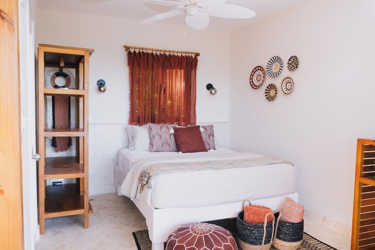 Un dormitorio con cama blanca, armario de madera, ventilador de techo, ventana con cortinas rojas y decoración en la pared.