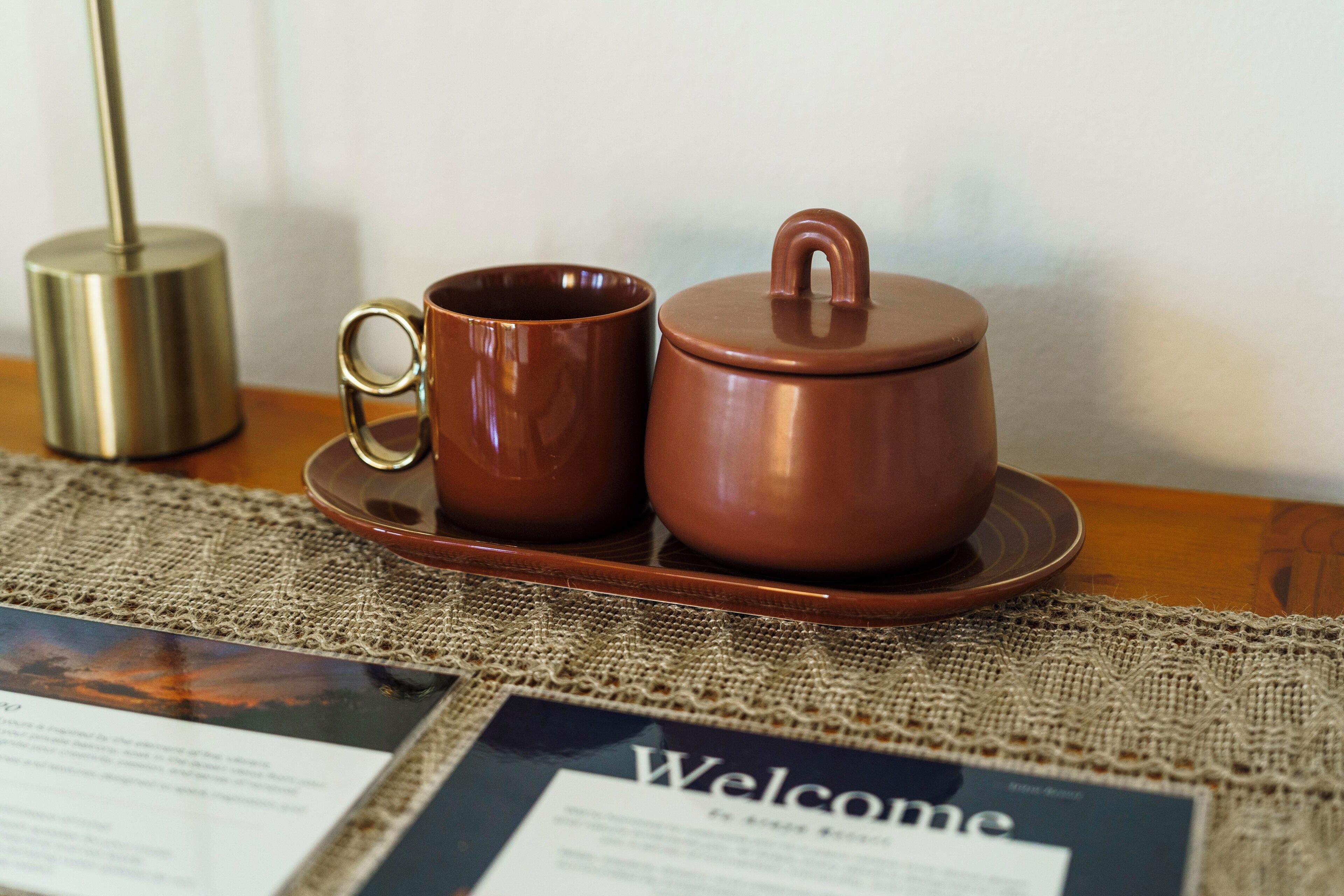 A tea set on a table with a burlap placemat and a welcome sign.