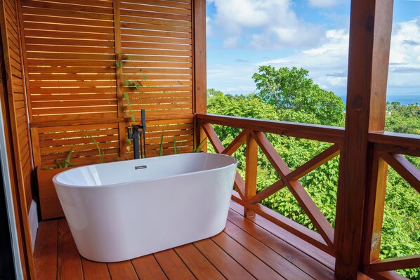 A white freestanding bathtub on a wooden deck with a view of lush greenery and the ocean. - Araya Resort (Samaná)