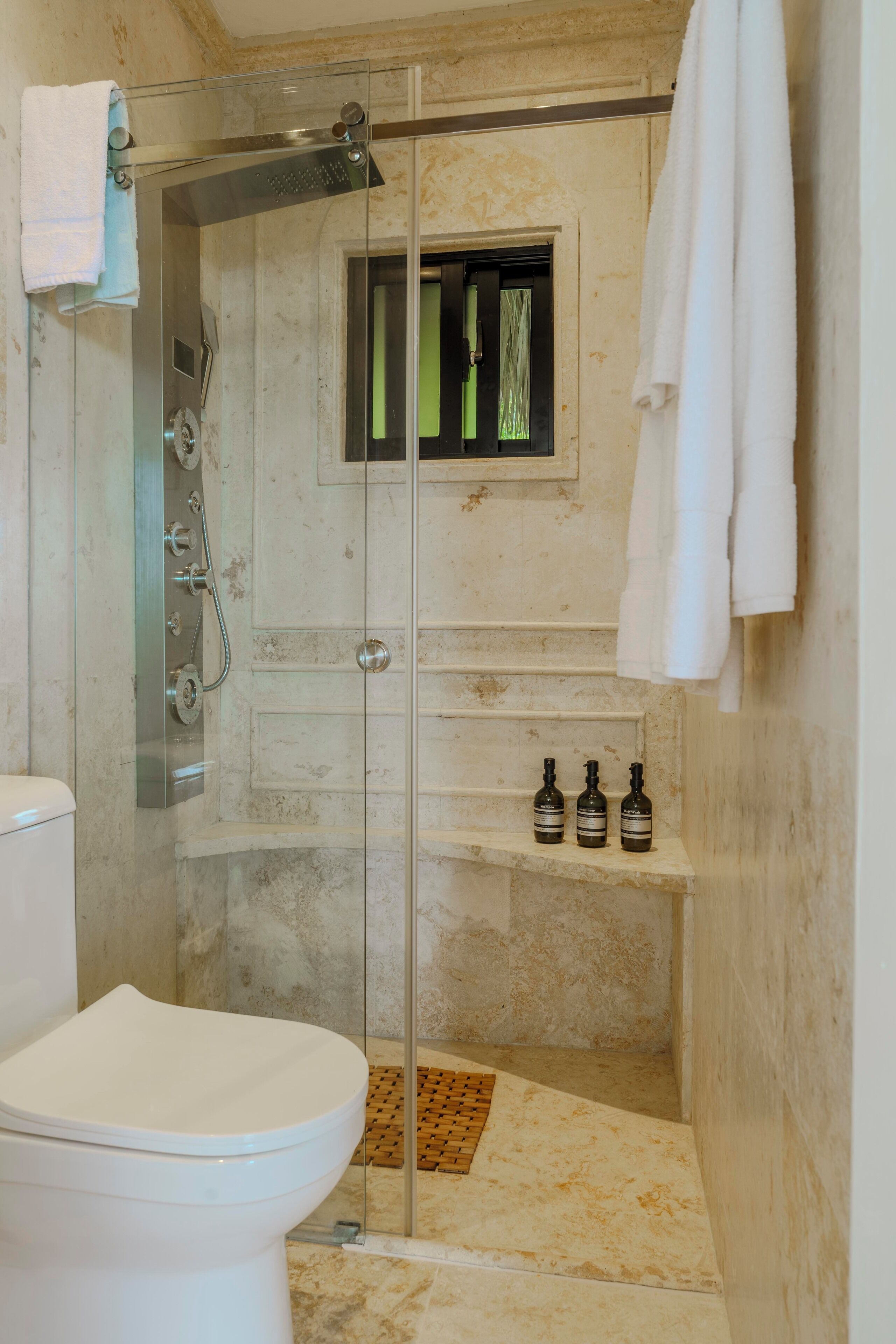 A bathroom with a glass-enclosed shower, a toilet, and a shelf with bottles.