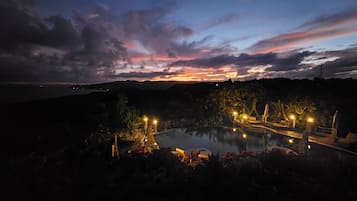 Vista nocturna de un resort con una gran piscina, iluminada por luces ambientales, rodeada de exuberante vegetación y un paisaje con atardecer de fondo.