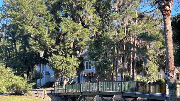 A park with a bridge over a pond, surrounded by trees and a house in the background. - Reduced fall rates! Evian.Sleeps 6 2BR 2.5 bath free pickleball, tennis & pool. (Hilton Head Island)