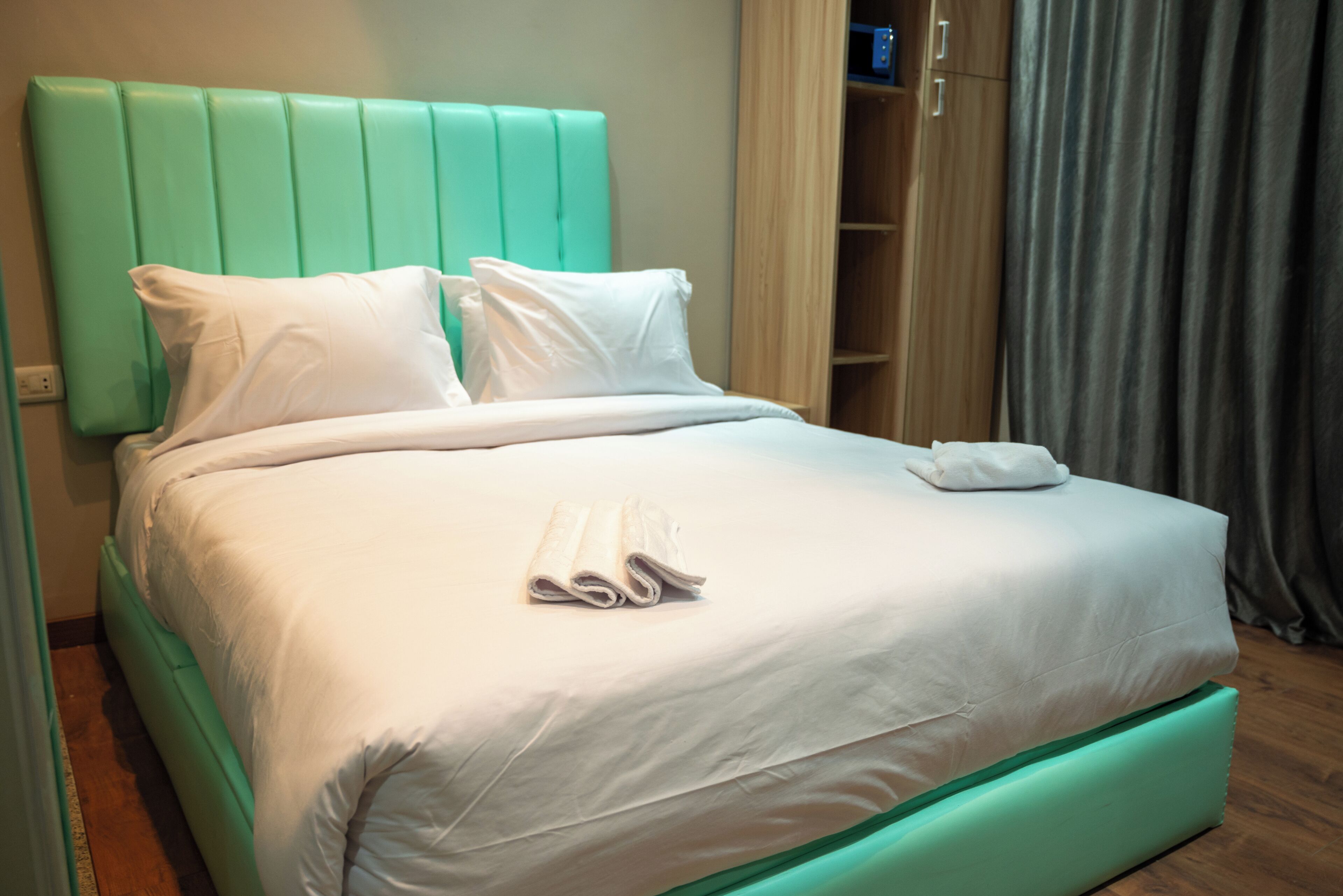 Una cama bien hecha con sábanas blancas y un cabecero verde azulado, un armario de madera y una cortina gris.