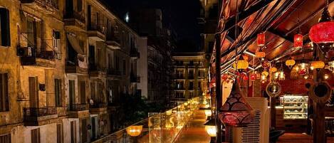 A nighttime view of a narrow street with buildings on either side, a reflective waterway, and outdoor seating with tables and chairs.