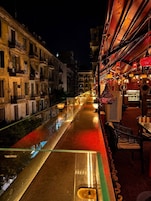Vista nocturna de una calle estrecha flanqueada por edificios, un cuerpo de agua reflectante y mesas y sillas para exteriores.
