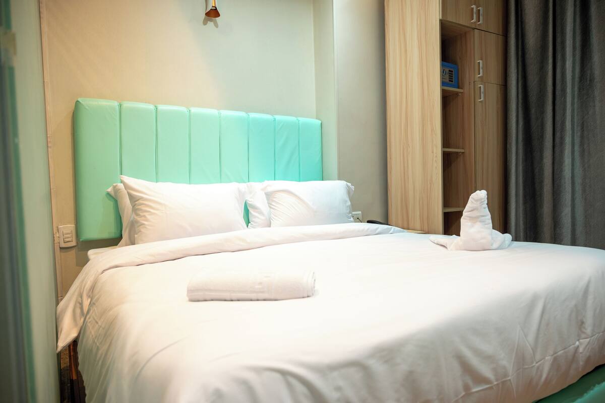 A neatly made bed with white linens and a teal headboard, a wooden wardrobe, and a folded towel on the bed.