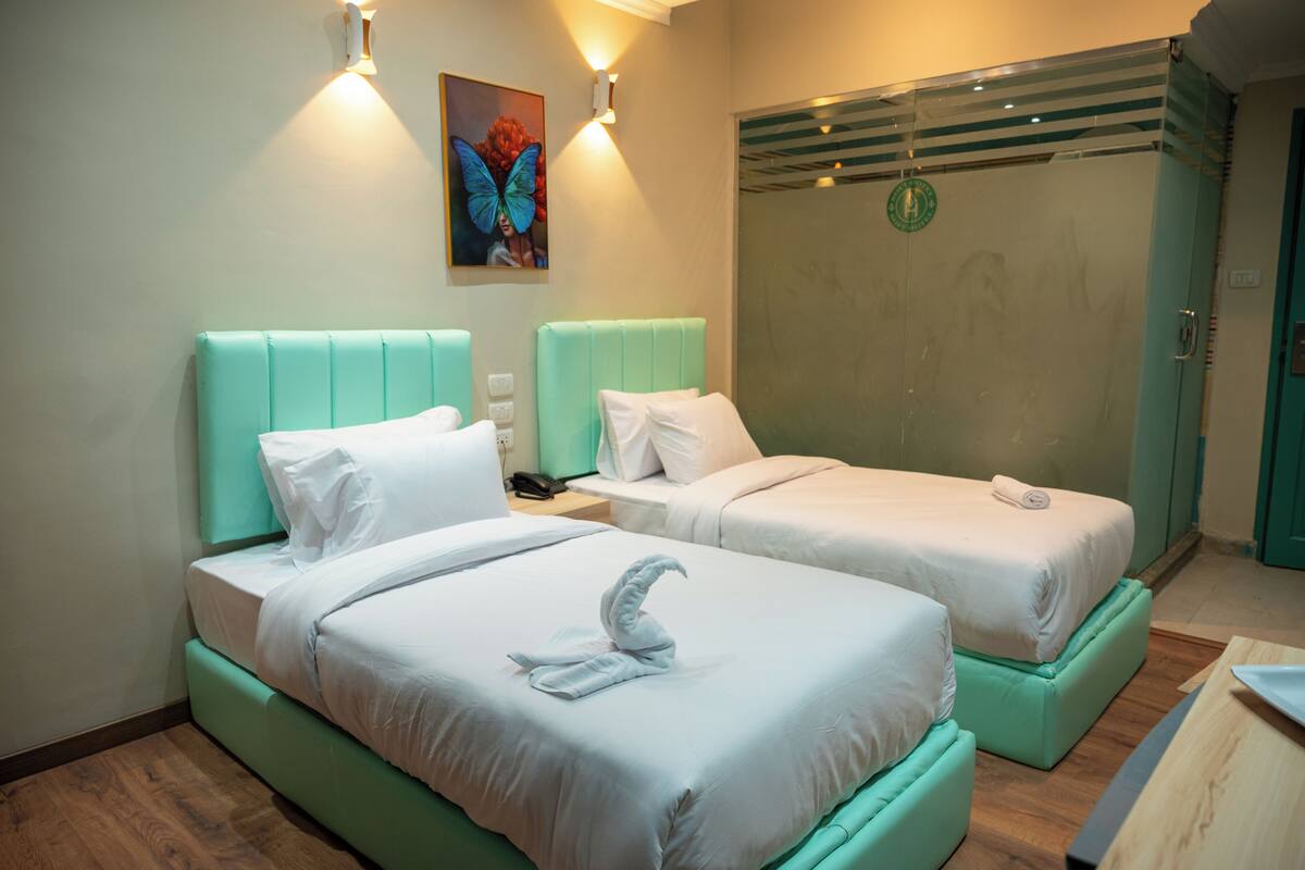 A hotel room with two beds, each with a teal headboard and white bedding. There is a painting of a butterfly on the wall, and a shower with a glass door.