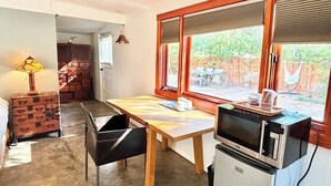 Cocina pequeña equipada con microondas, una mesa y una silla. Se ve una ventana con persianas, una lámpara y una maleta.