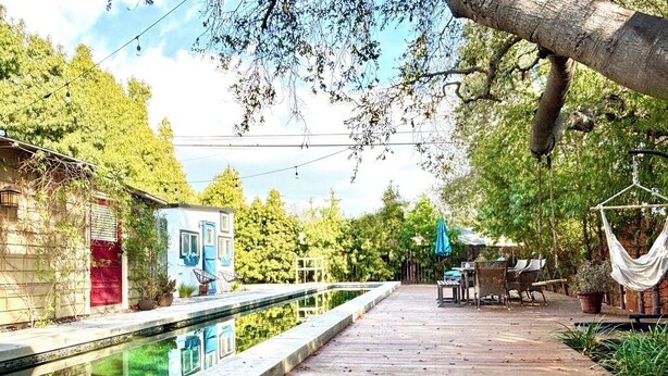 Una terraza de madera con una piscina, rodeada de follaje y un pequeño edificio con puerta roja.