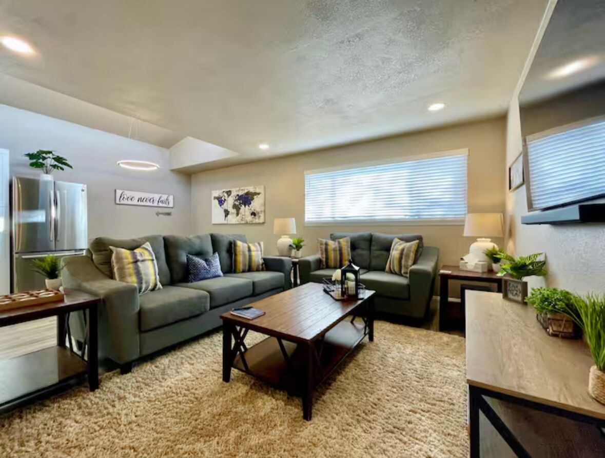 A living room with two sofas, a coffee table, and a TV. There is a plant on the left and a window with blinds on the right.