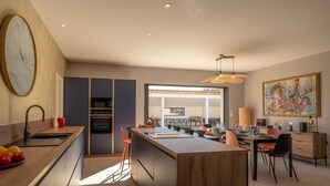 A modern kitchen with a large clock, a dining area with a view, and a painting on the wall.